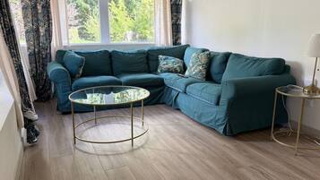 A corner living room with a teal sofa, a glass coffee table, and a lamp on a side table.