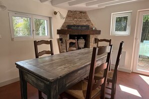 A dining room with a rustic brick fireplace, a wooden table, and two windows.