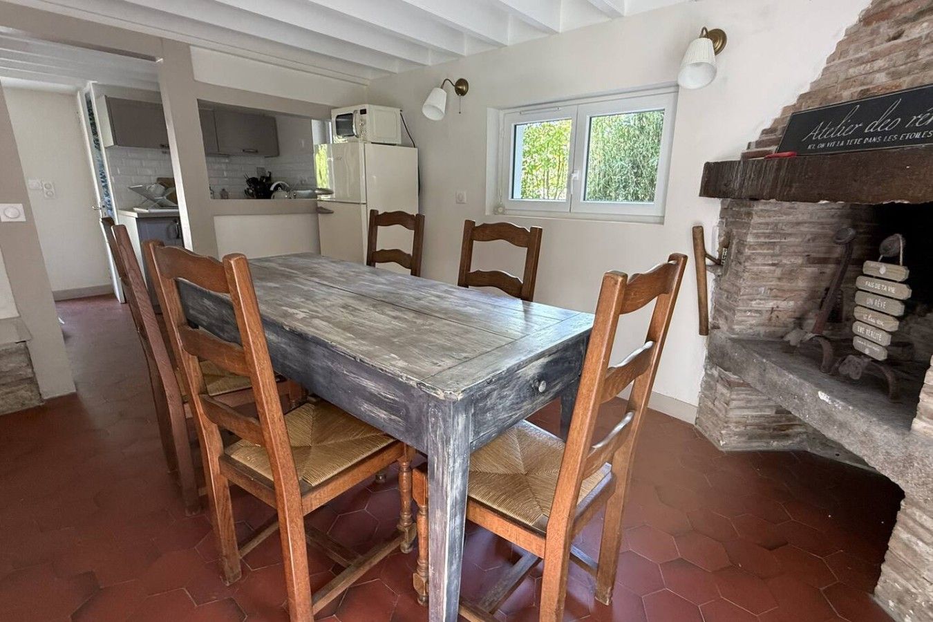 A dining area with a rustic wooden table and chairs, a kitchen in the background, and a brick fireplace.