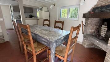 A dining area with a rustic wooden table and chairs, a kitchen in the background, and a brick fireplace.