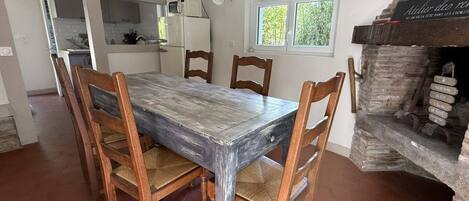 Un coin repas avec une table et des chaises en bois rustique, une cuisine en arrière-plan et une cheminée en briques.