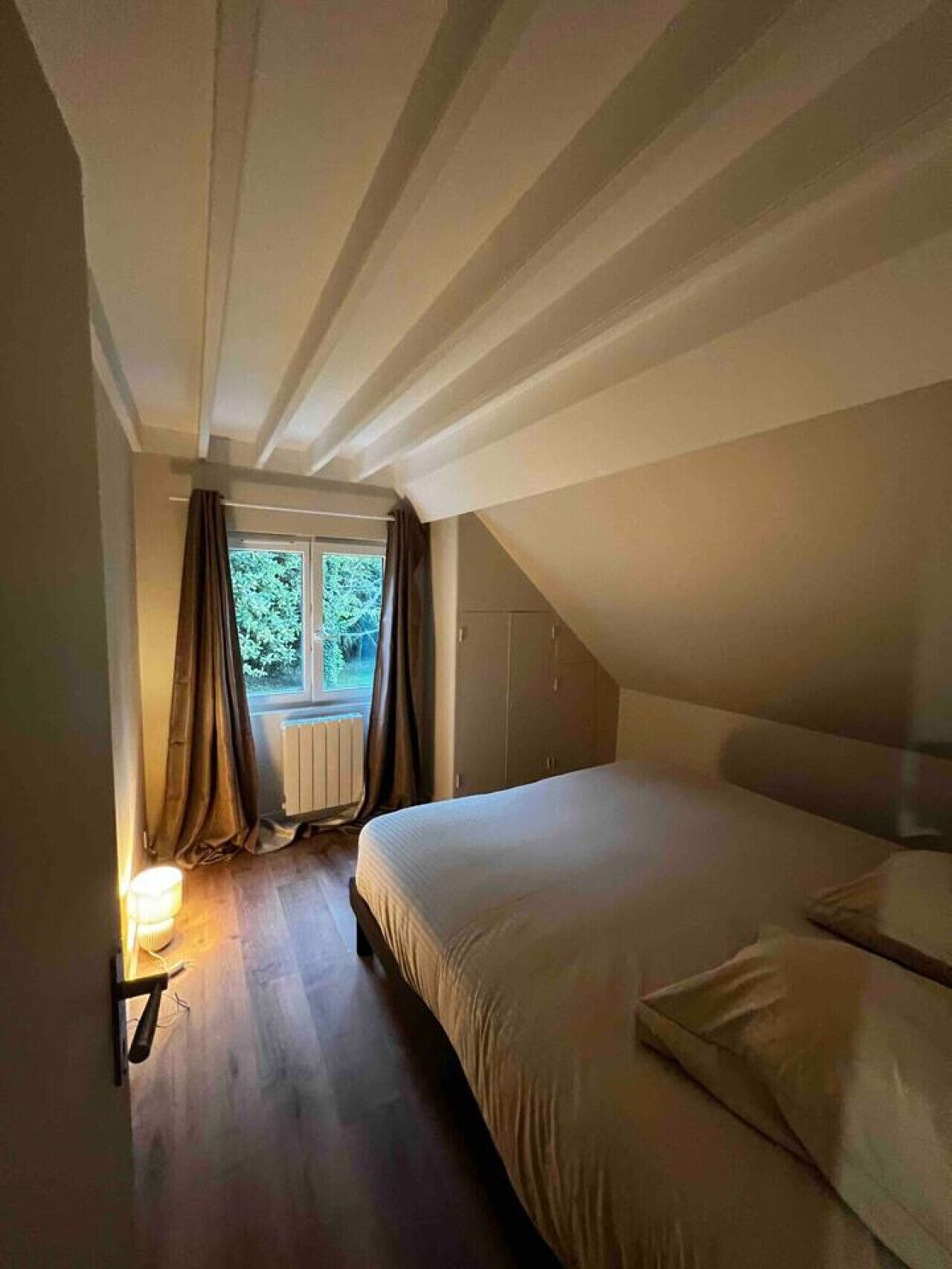 Une chambre avec un plafond en pente, une fenêtre avec des rideaux, un lit et une table de chevet avec une lampe.