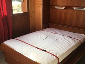 Una cama individual con colchón blanco y correas rojas, paredes de madera, una ventana con cortinas y un armario de madera.