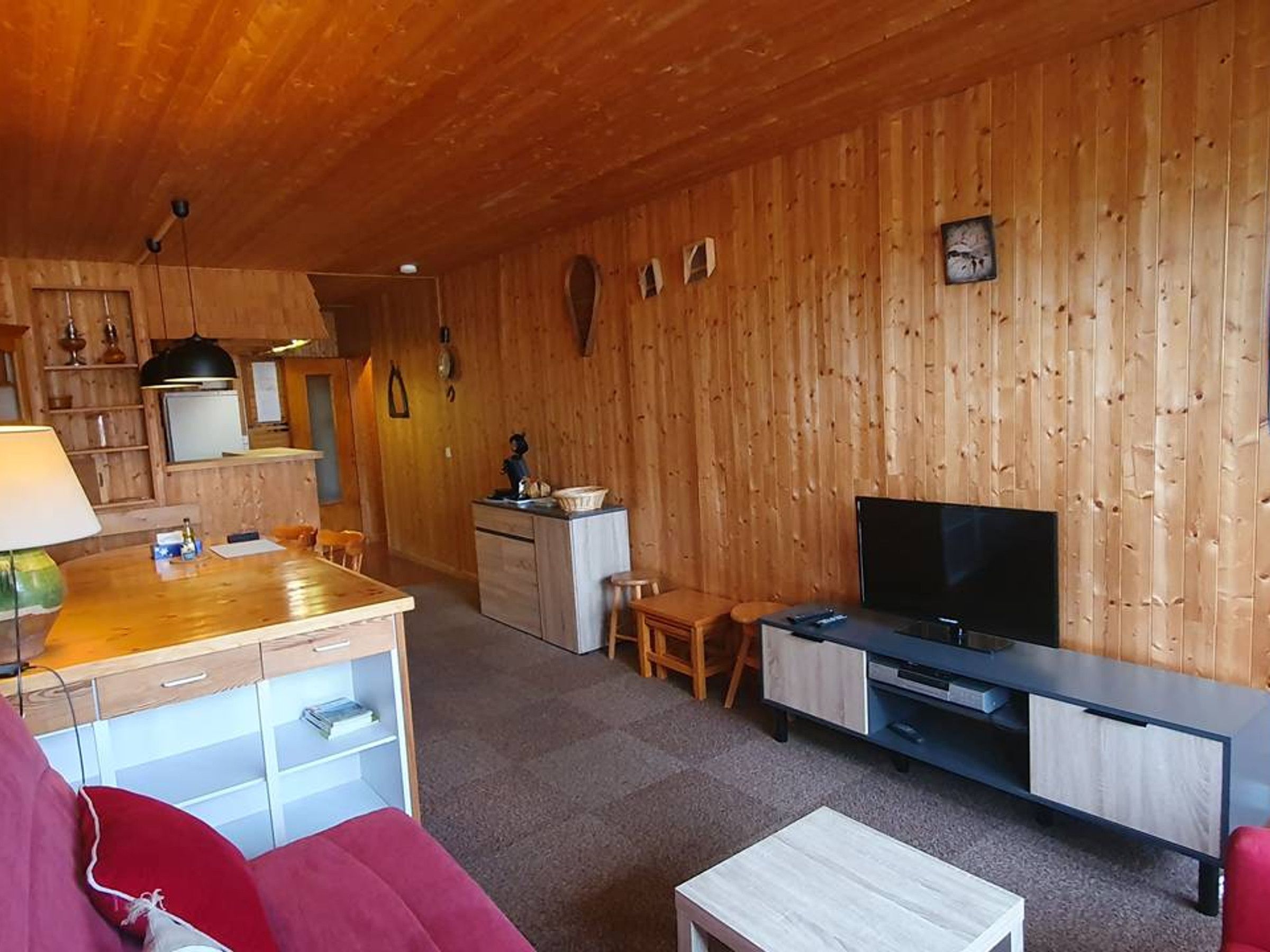 A cozy living room with a TV, wooden walls, a kitchen area with a fridge, and a dining table.