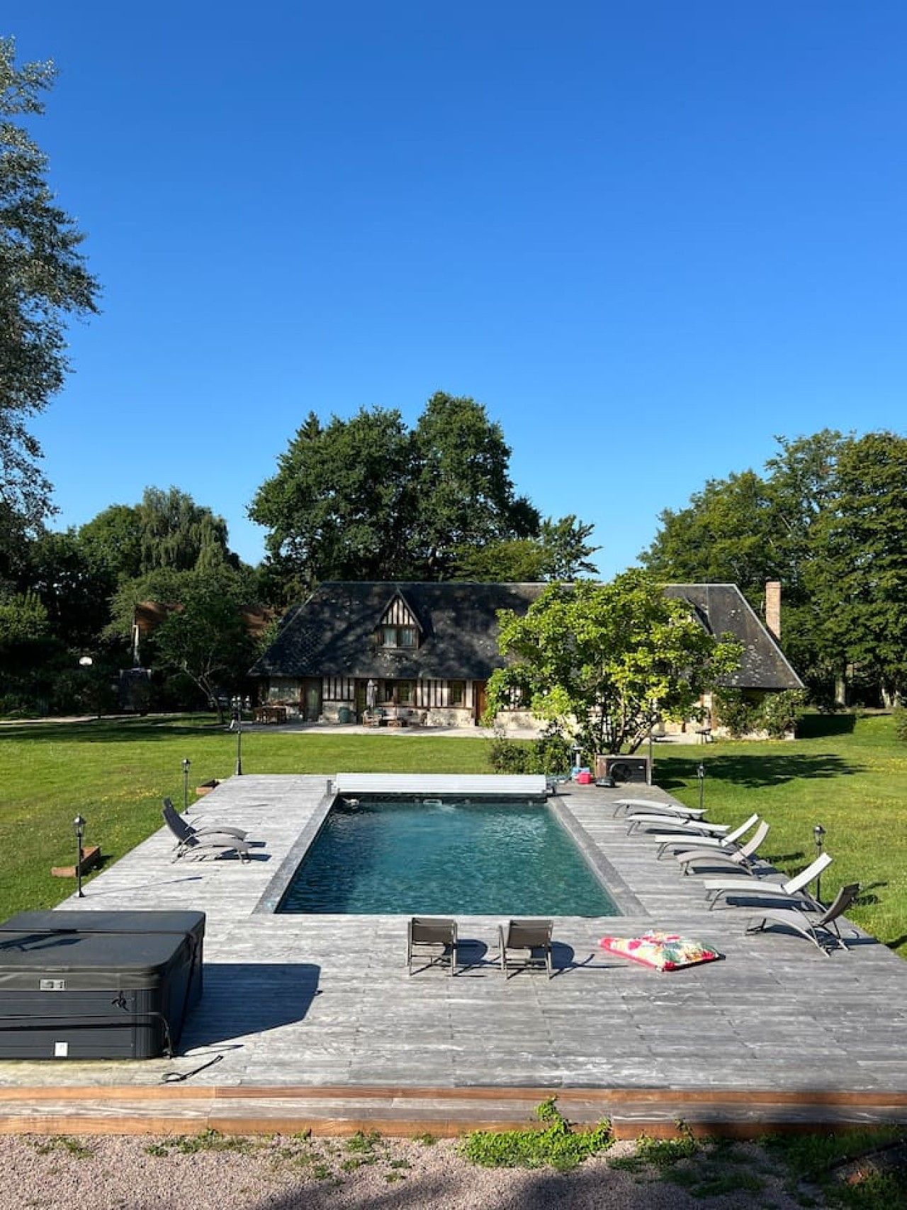 Una piscina con lettini e una valigia su un solarium, circondata da alberi e con una casa sullo sfondo.