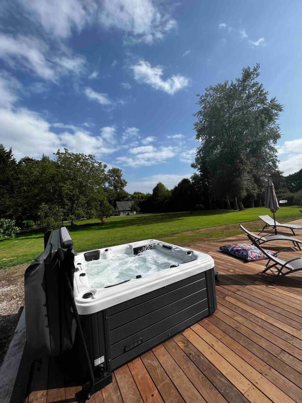 A hot tub on a wooden deck with lounge chairs and a covered umbrella, surrounded by a grassy yard and trees under a clear sky.