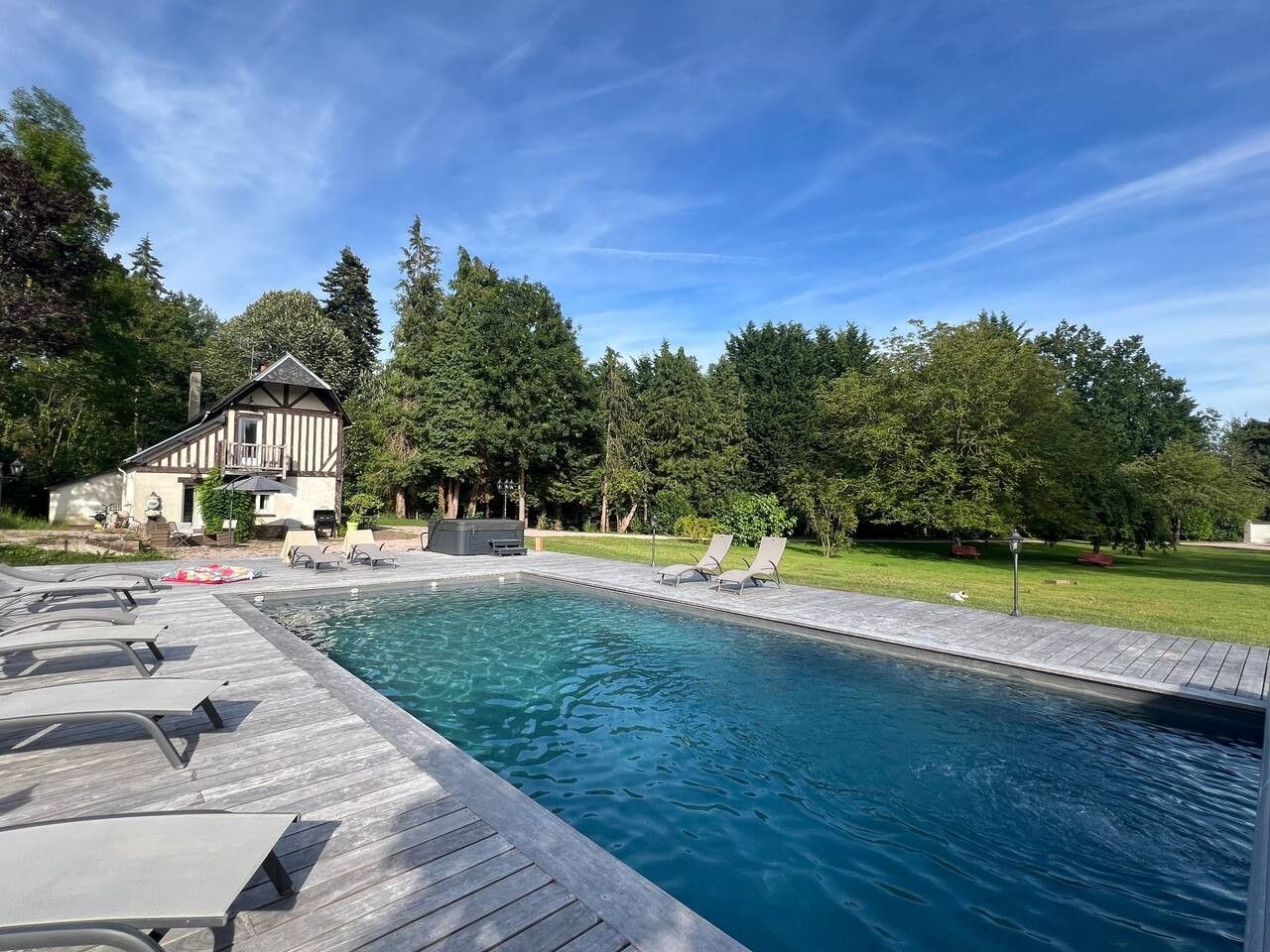 Una piscina con muelle de madera y sillas de descanso, rodeada de árboles y una casa.