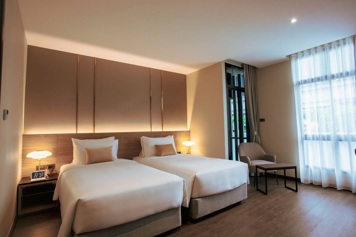 Quarto de hotel com duas camas, uma mesa de cabeceira com abajur, uma cadeira e uma janela grande com cortinas.