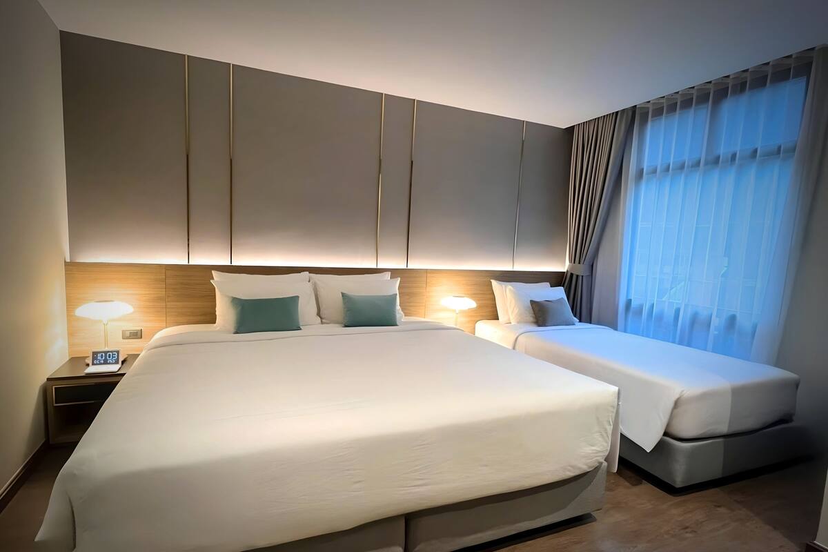 Quarto de hotel moderno com uma cama grande, uma cama menor e uma mesa de cabeceira com abajur.