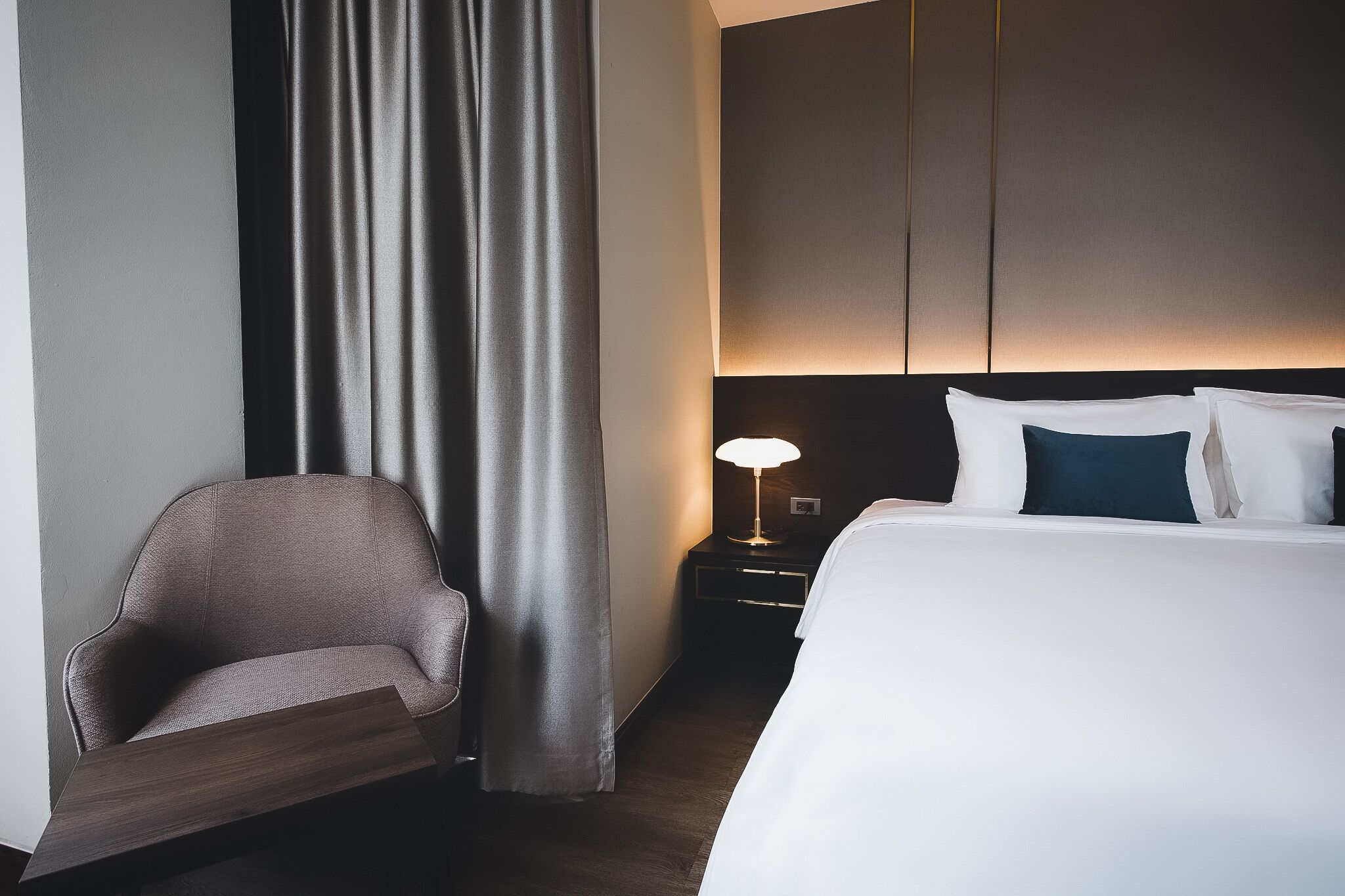A hotel room with a bed, a chair, a nightstand with a lamp, and a curtain.