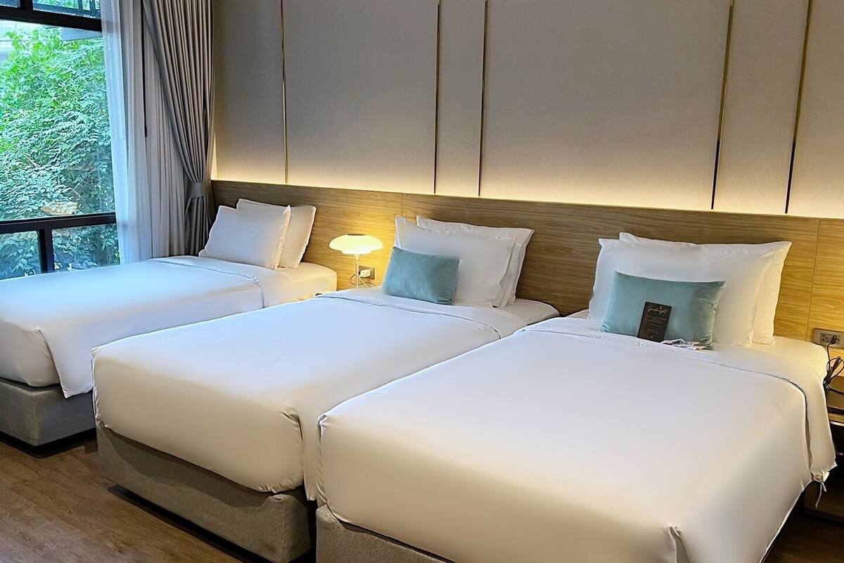 Quarto de hotel com três camas, uma janela grande com cortinas e vista para o verde lá fora.