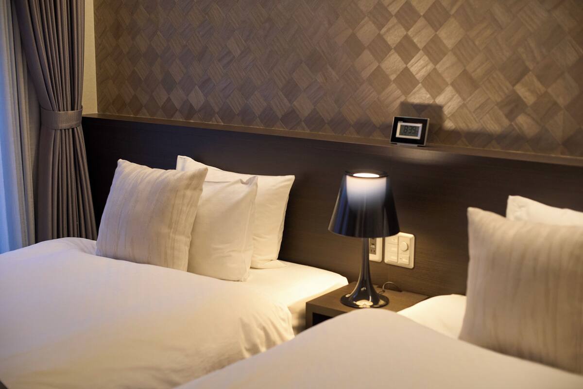 Una habitación de hotel con cama, almohadas, mesita de noche, lámpara y un reloj en la pared.