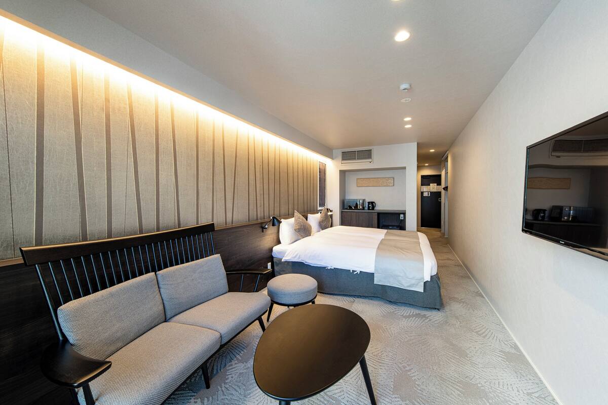 Una habitación de hotel moderna con un sofá, una mesita redonda pequeña, una cama con edredón blanco y un televisor montado en la pared.