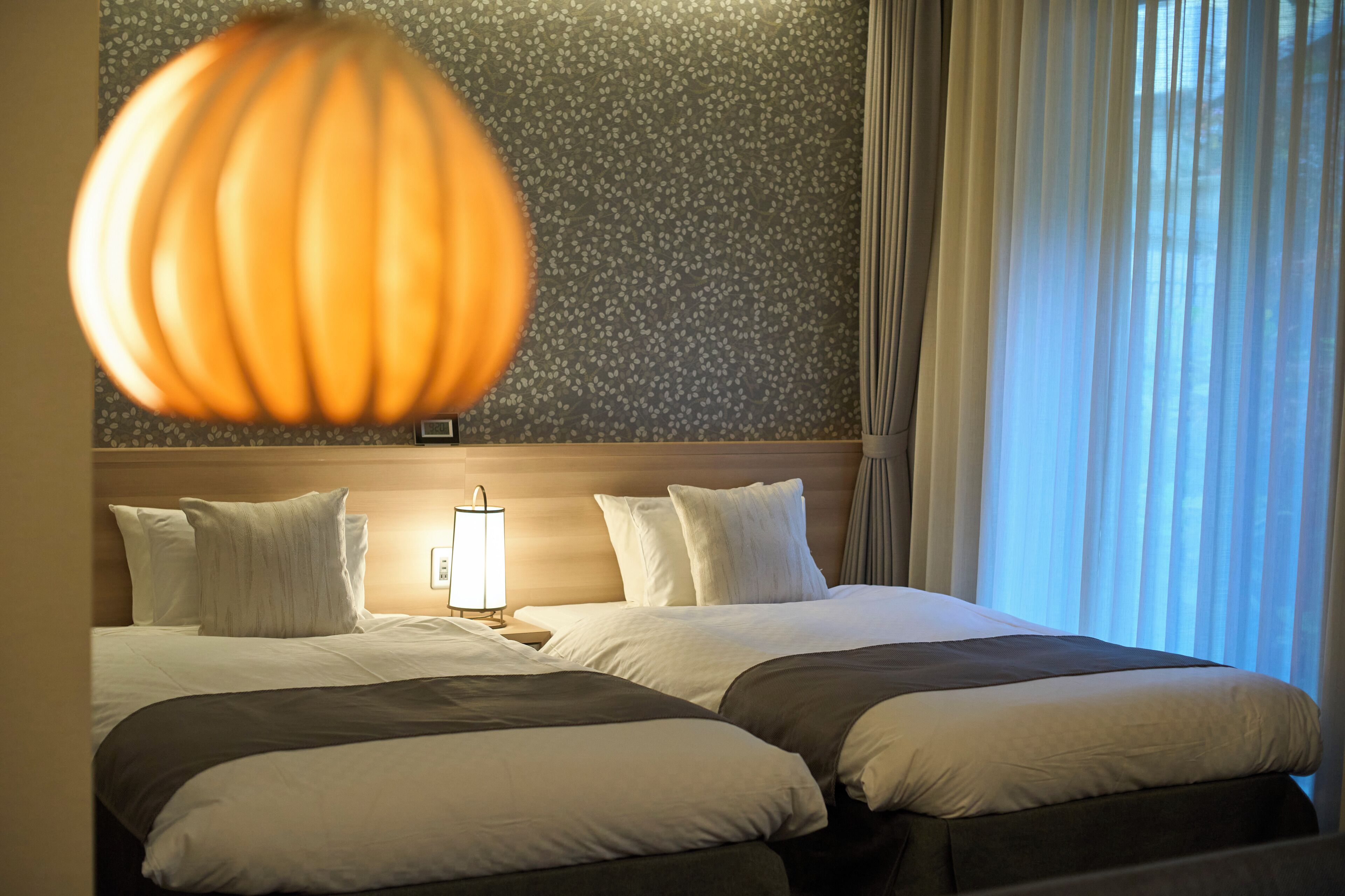A hotel room with two beds, a nightstand with a lamp, and a window with curtains.