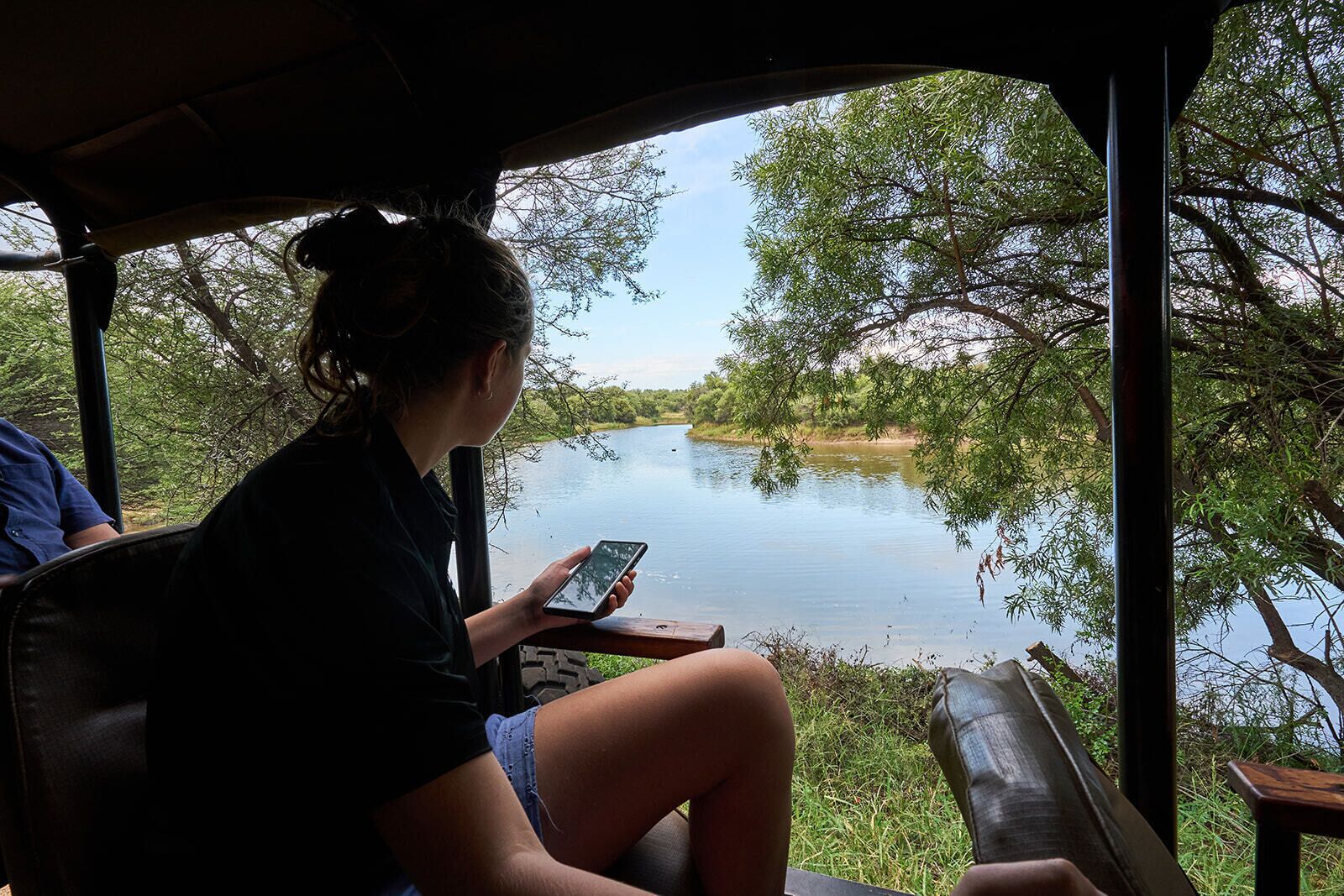 A person seated inside a vehicle, holding a smartphone, with a view of a calm river and greenery outside.