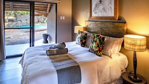 A well-lit bedroom with a large bed, a view of an outdoor area through a sliding door, a framed picture on the wall, and a bedside lamp.