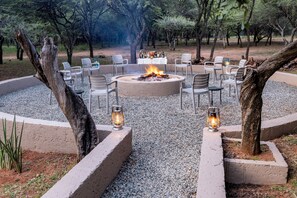 A circular outdoor seating area with a fire pit, surrounded by chairs and tables, set in a natural environment with trees and gravel.