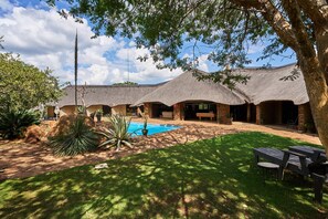 A thatched-roof building with a swimming pool, surrounded by greenery and a lawn. - Sable Ranch Bush Lodge  (Brits)