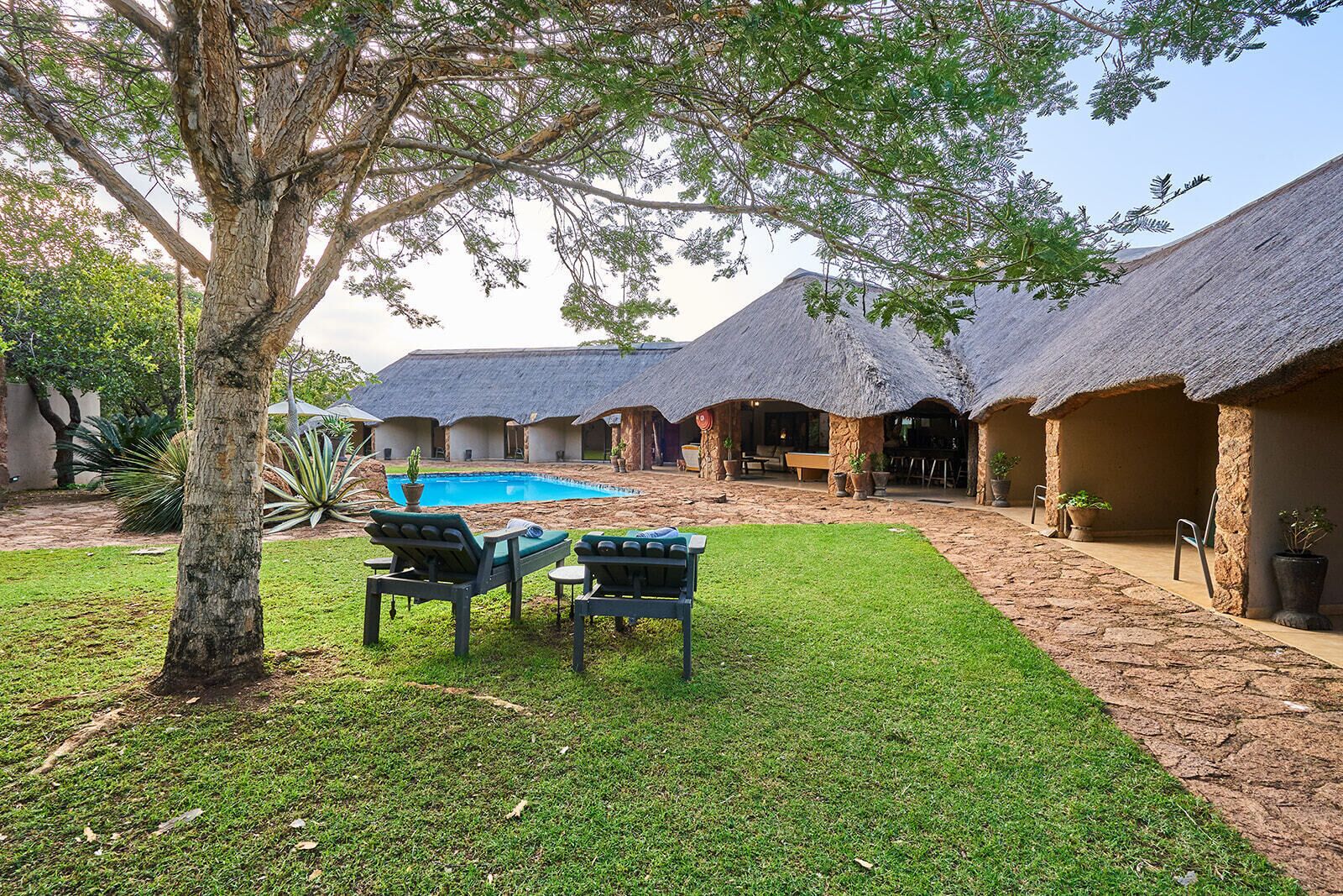 A pool area with lounge chairs, a thatched-roof building, and a tree.