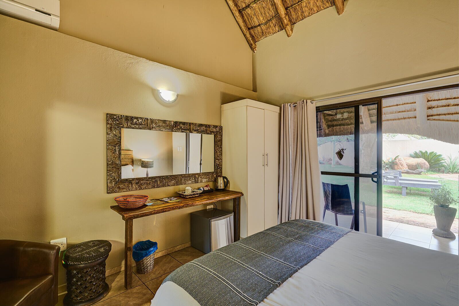 Une chambre à coucher comprenant un lit, un bureau avec un miroir, une armoire, une poubelle et une vue sur l’extérieur.