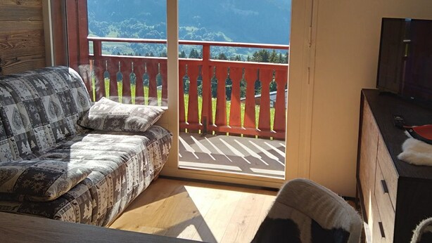 A room with a sofa, a TV, and a view of mountains through a balcony with a red railing. - Cozy Studio for 4, Near Cernix Center and Slopes (Crest-Voland)