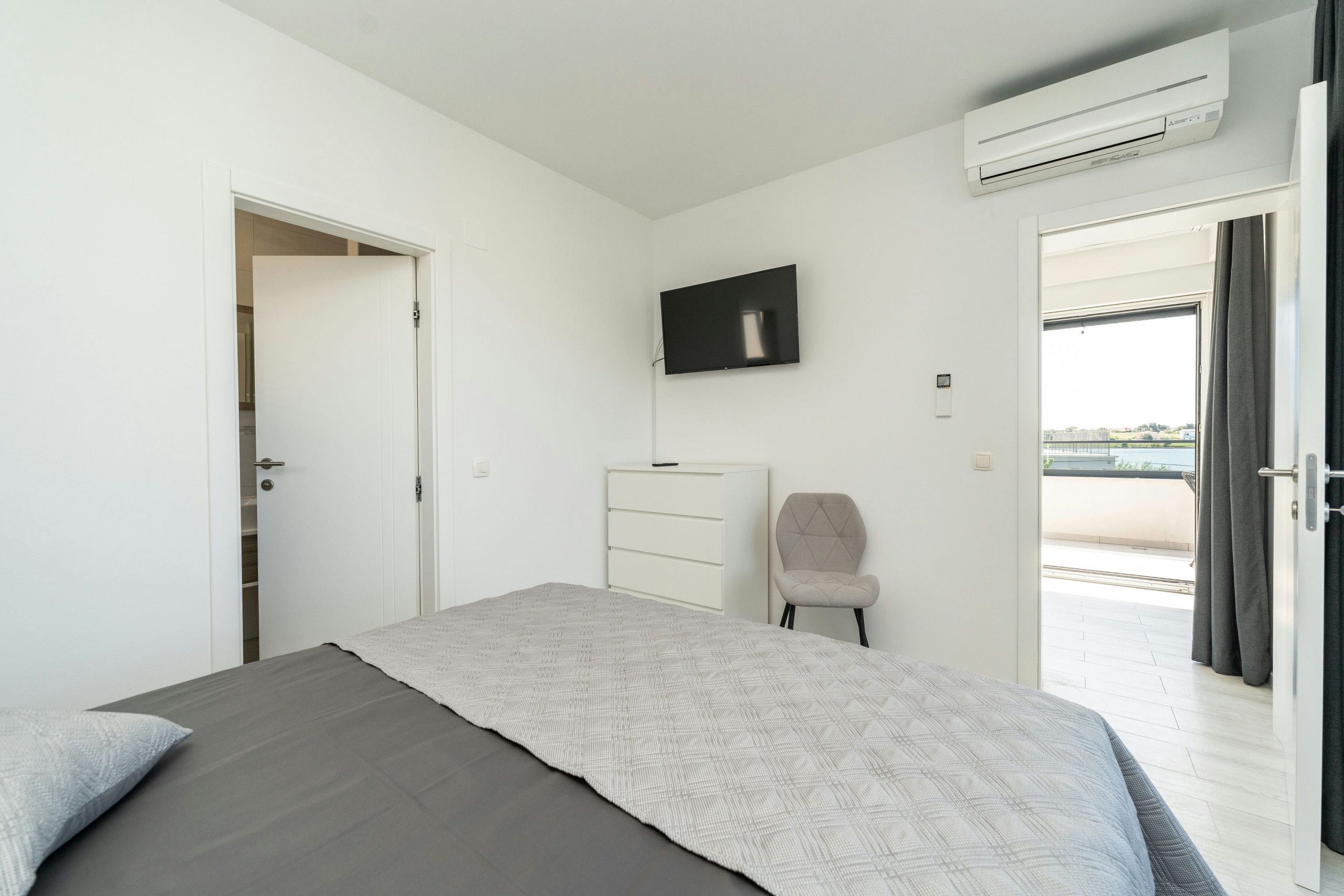Un dormitorio con cama, televisor montado en la pared, una silla, cómoda y equipo de aire acondicionado.
