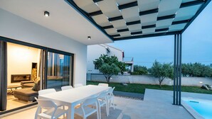 Een modern terras buiten met een witte tafel en stoelen, een glazen deur naar een leefruimte en een zwembad.