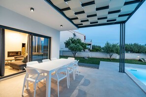 Une terrasse extérieure moderne avec une table et des chaises blanches, une porte vitrée donnant sur un espace de vie et une piscine.