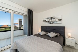Une chambre moderne avec un grand lit, une table de chevet, une lampe fixée au mur et une vue sur les bâtiments à travers une porte-fenêtre coulissante.