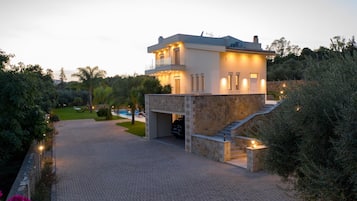Uma casa moderna de dois andares com fachada de pedra, uma entrada de automóveis que leva a uma garagem e um jardim ajardinado com piscina.