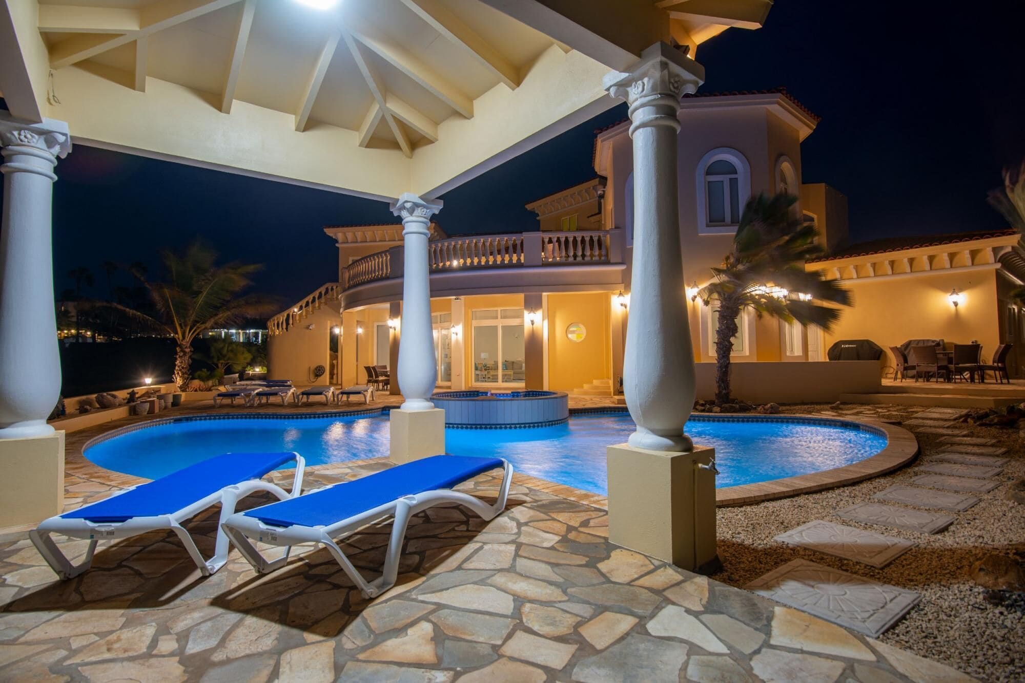 Un área junto a la piscina con sillones reclinables, piscina y un edificio con columnas y balcón.