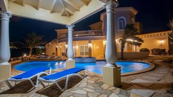 Un área junto a la piscina con sillones reclinables, piscina y un edificio con columnas y balcón.