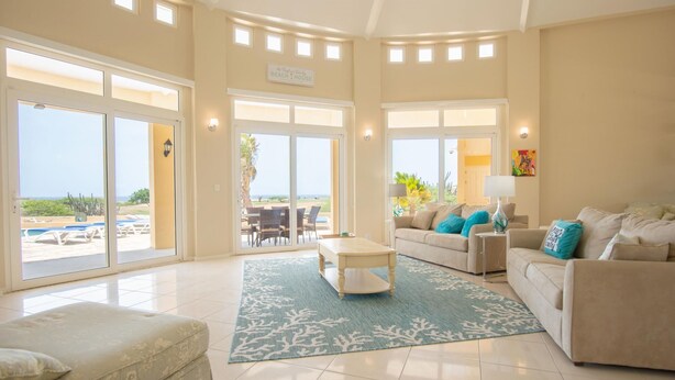 A spacious living room with a large sliding glass door leading to a balcony, a beige sofa set, a coffee table, and a patterned rug. - This One of a Kind Amazing Aruba Beach House (Noord)