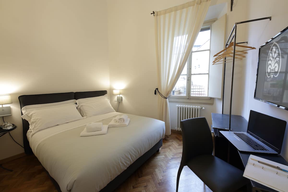 Uma cama bem arrumada com lençóis brancos, um laptop numa mesa e uma cadeira.