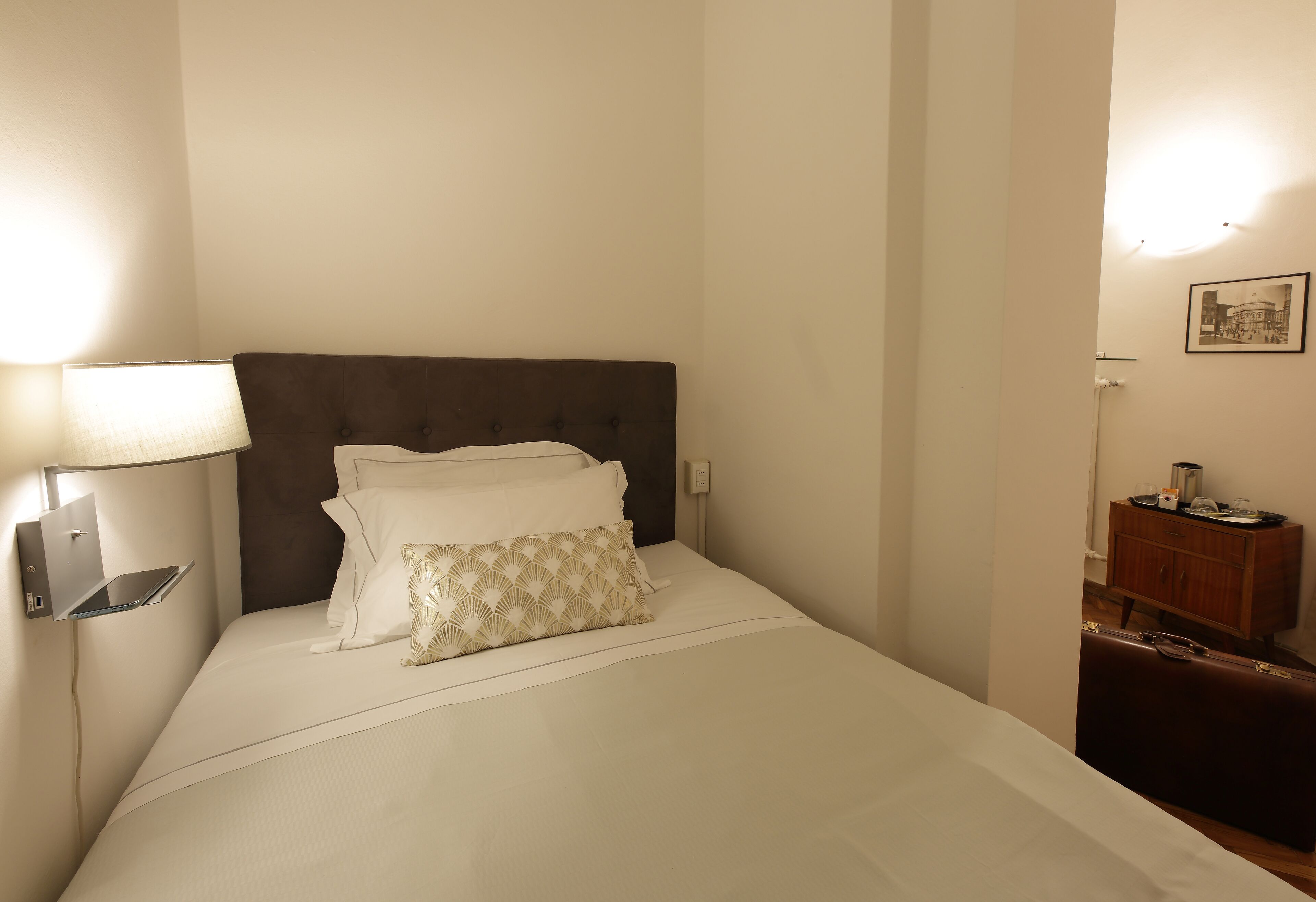 Un lit bien fait, avec du linge de lit blanc et un coussin à motifs, une lampe de chevet et une petite table de nuit en bois sur laquelle se trouve une autre lampe.