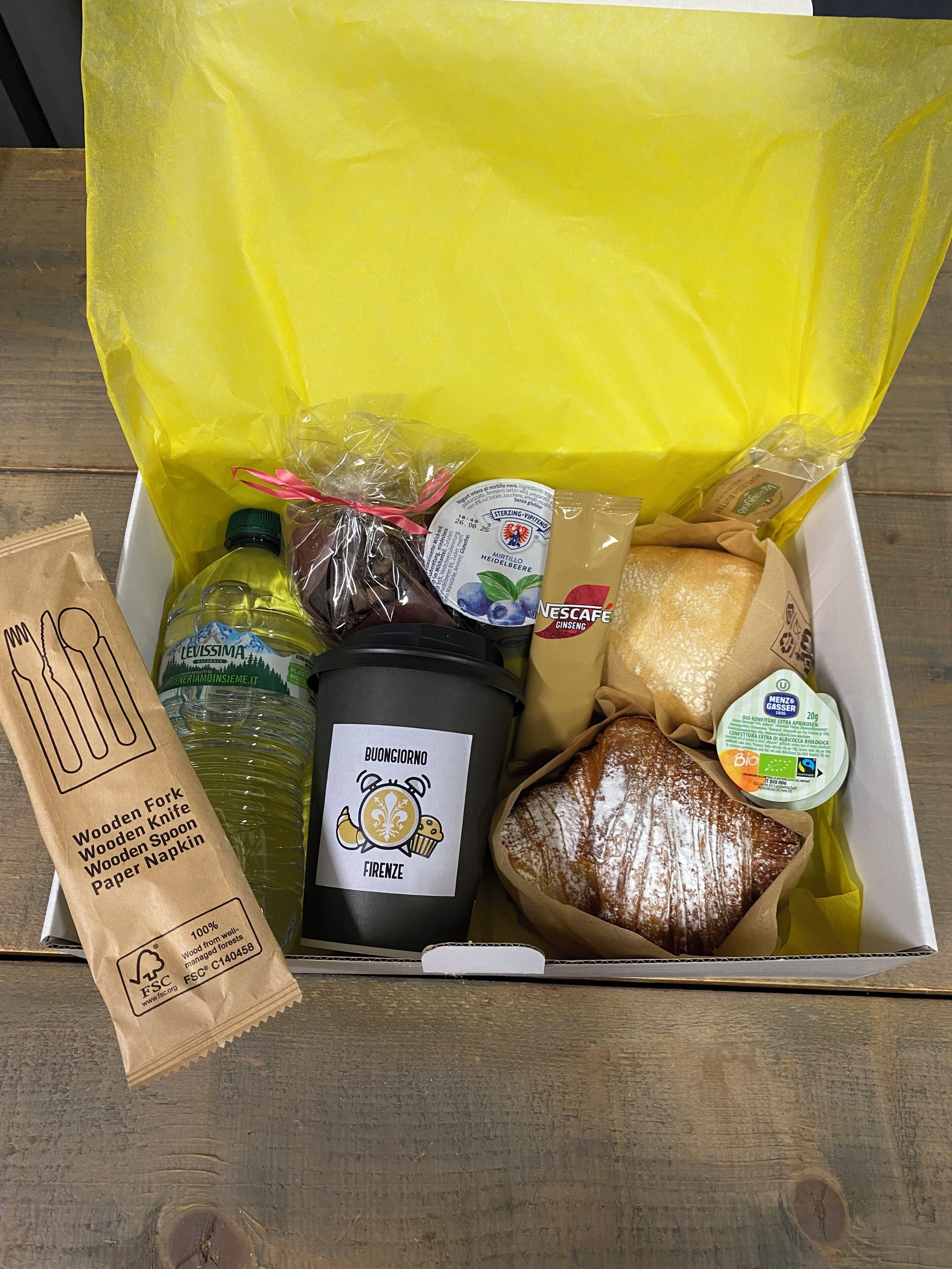A gift box containing various food items including a bottle of olive oil, a jar of pesto, a loaf of bread, and a package of cheese.