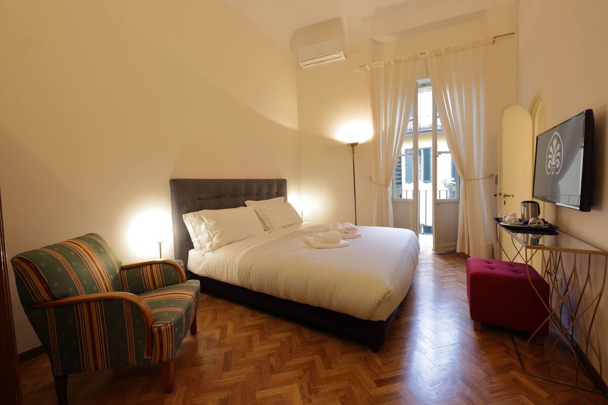 Quarto com uma cama, uma cadeira, uma mesinha e uma televisão na parede.