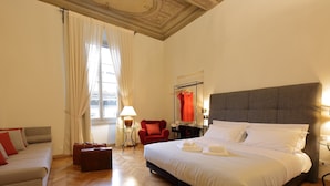 Una habitación de hotel amplia con una cama grande, una zona de estar con sofá y sillón, una mesita con lámpara y un ventanal con cortinas.