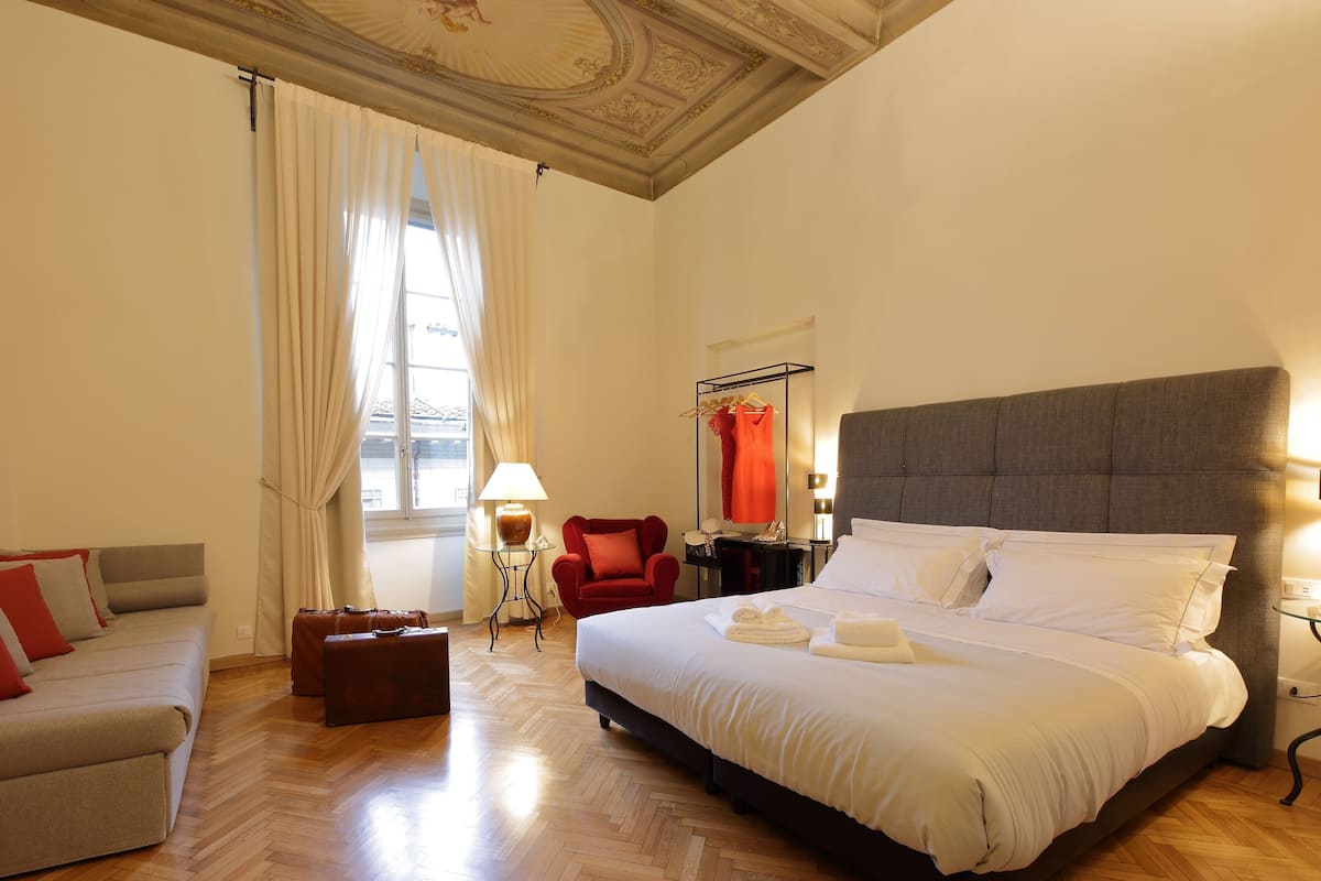 Um quarto de hotel espaçoso, com uma cama grande, área de estar com sofá e poltrona, pequena mesa com abajur e janela com cortinas.