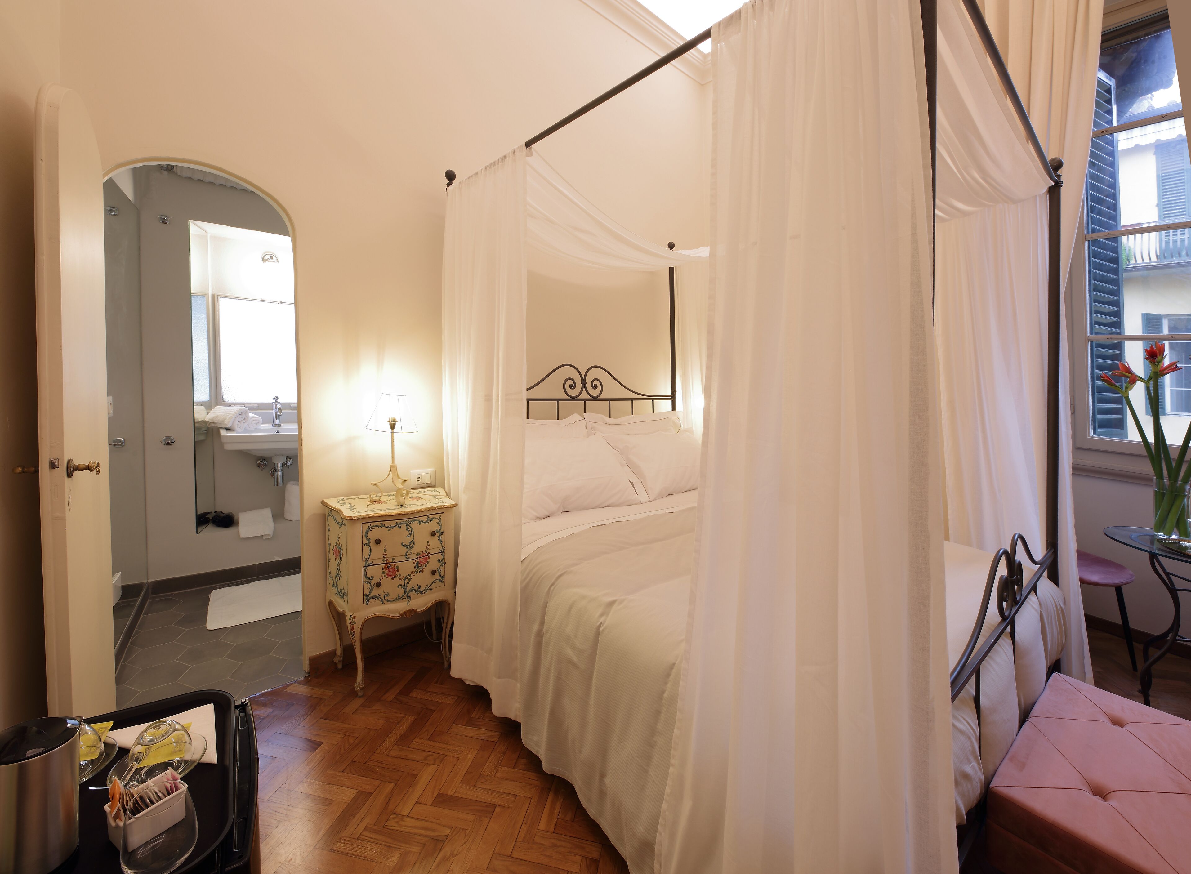 A bedroom with a four-poster bed, a bedside table with a lamp, a small table with a vase of flowers, and a view of the outside through a window.