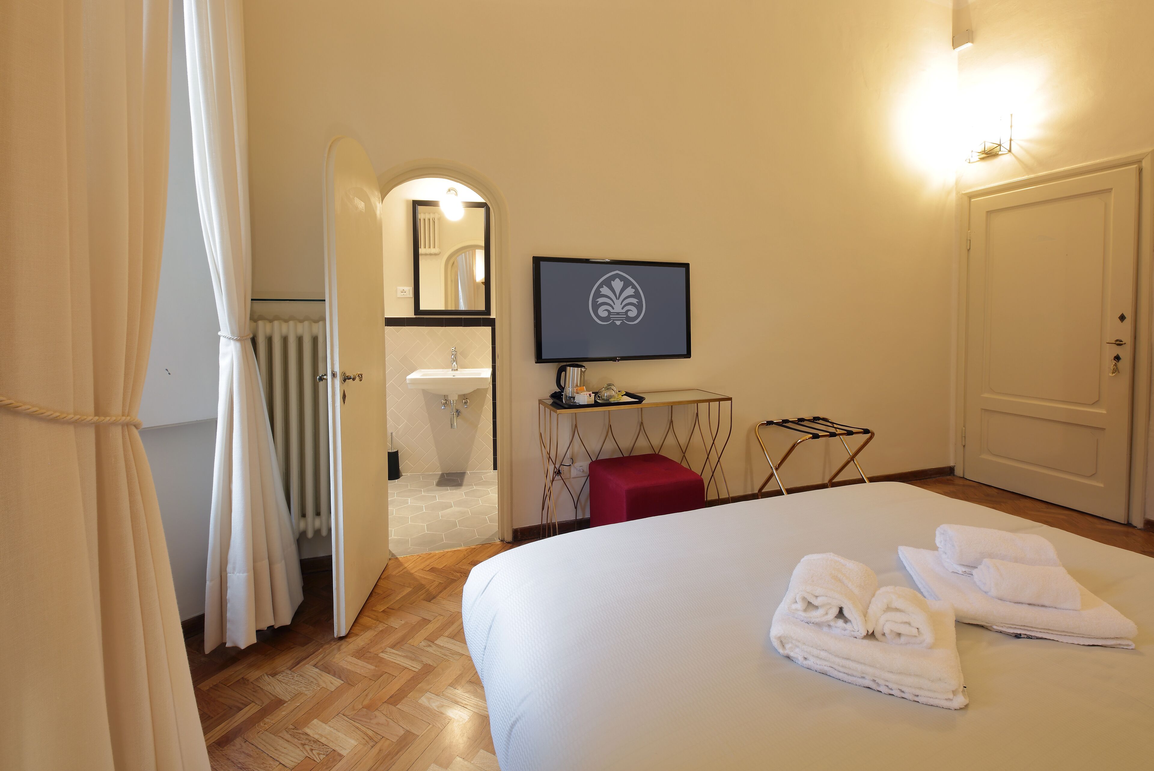 Une chambre d’hôtel avec un lit, une télévision, un miroir, un tabouret rouge et une salle de bain visible à travers une porte ouverte.