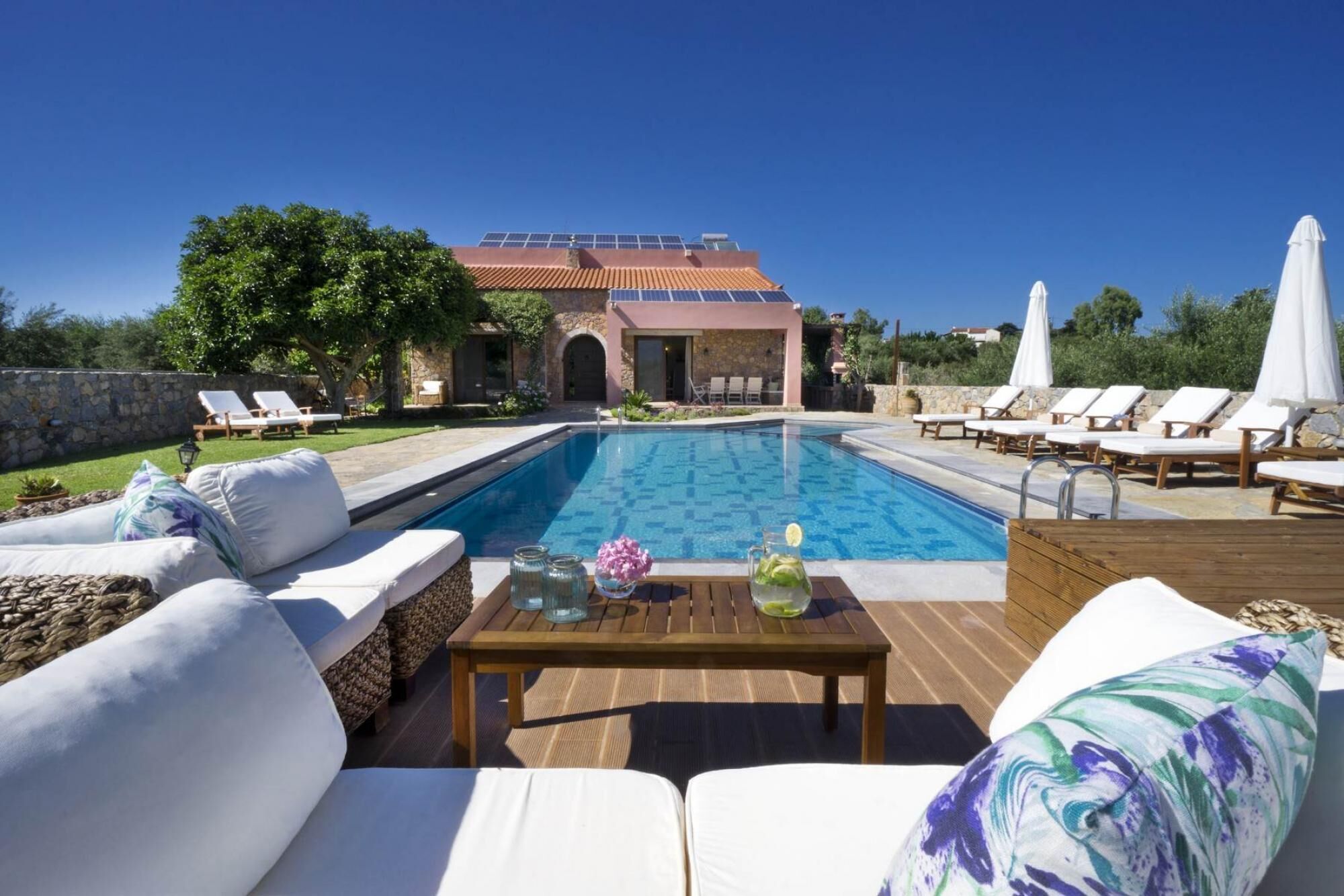 A pool area with lounge chairs, a wooden table with drinks, and a building with solar panels in the background.
