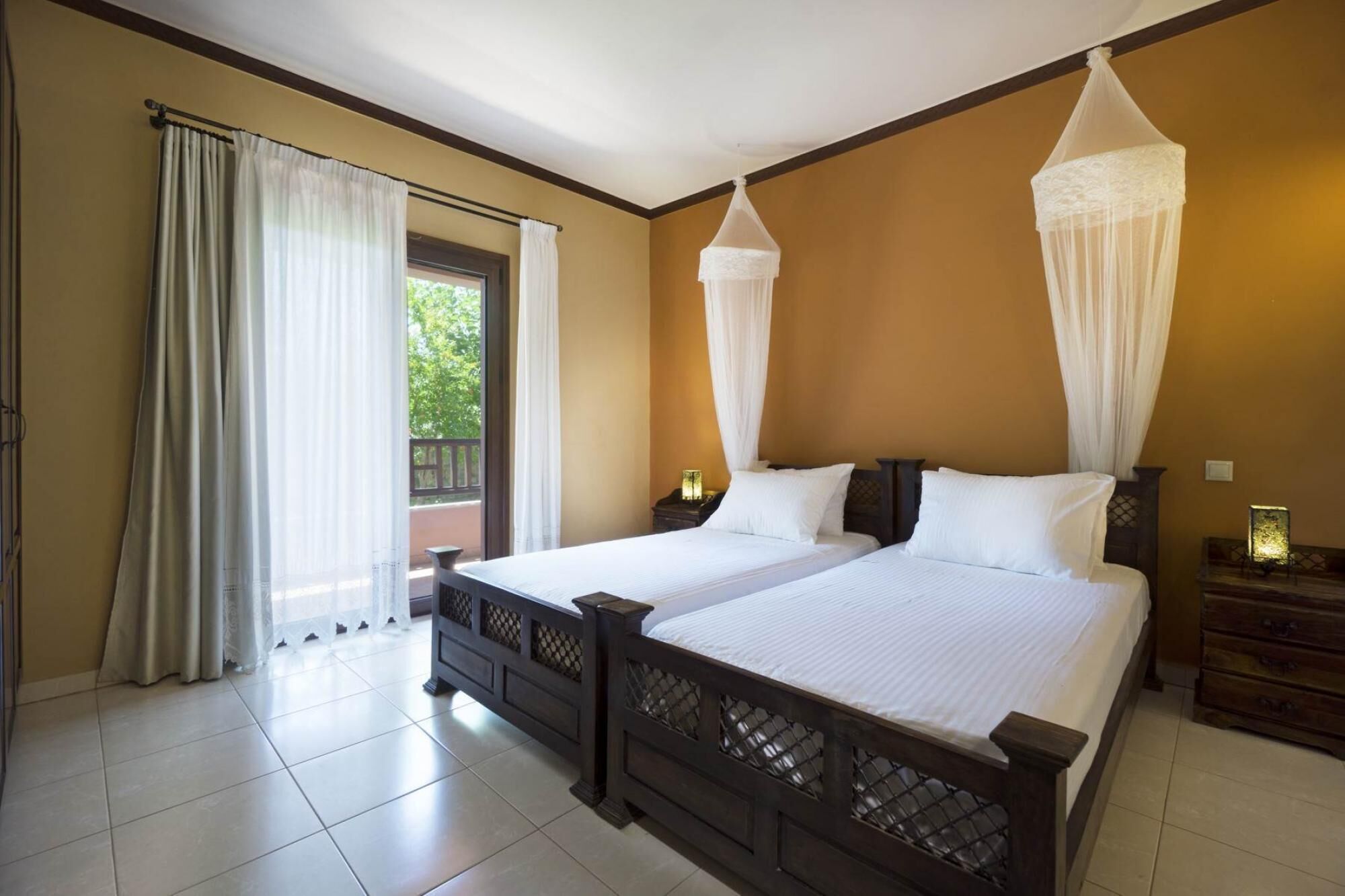 A double bed with a wooden frame and white bedding, a nightstand with a lamp, and a window with sheer curtains.