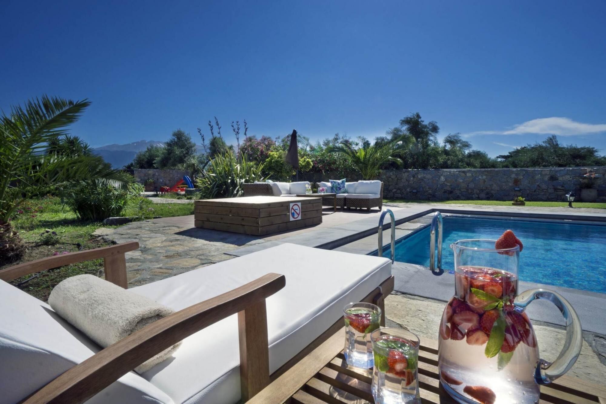 A poolside area with lounge chairs, a wooden table with drinks, and a stone wall in the background.