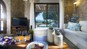 A cozy living room with a stone wall, a television, a sofa, a dining table with food and drinks, and a view of the outdoors. - Nafsika Villa in Litsarda (Apokoronas)
