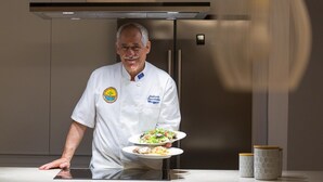 En kock i vit uniform med en logotyp, som håller i tallrikar med mat i ett modernt kök med rostfria köksredskap och en köksfläkt.