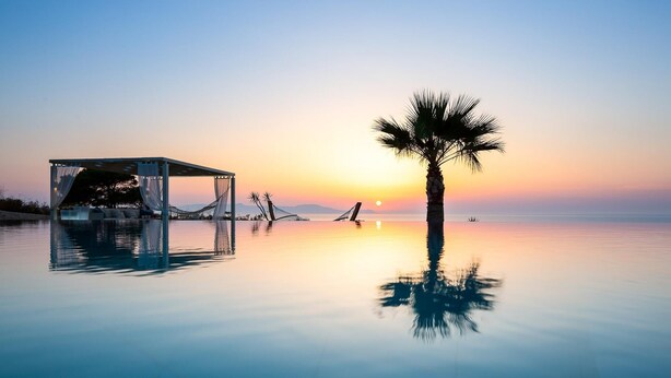 En lugn och vacker infinitypool som speglar en solnedgång, ett palmträd och en möblerad sittplats med utsikt över havet.