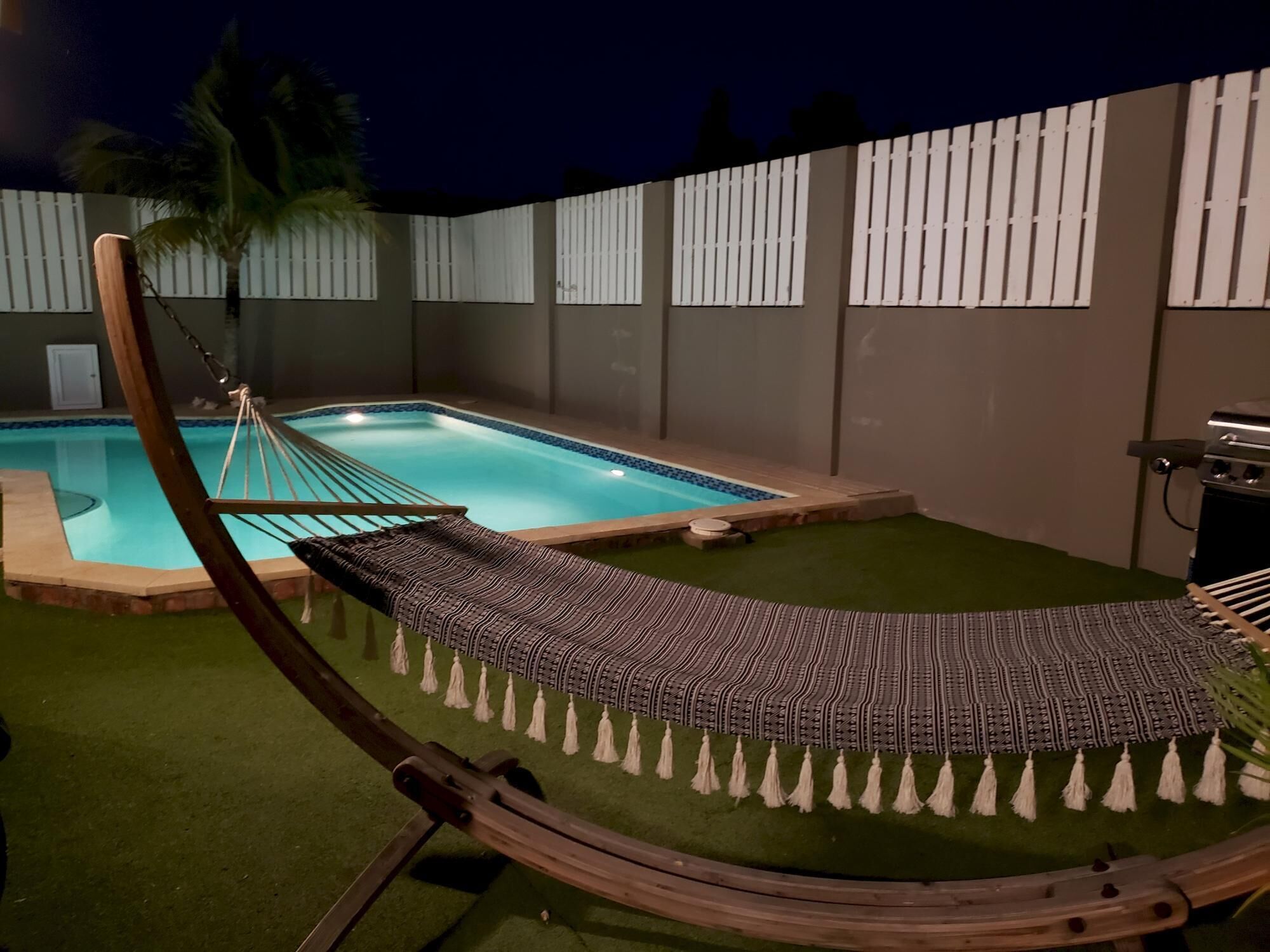 Una hamaca junto a una piscina, con una valla y una parrilla al fondo.