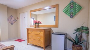 A hotel room with a wooden dresser, a small refrigerator, a mirror, a potted plant, and a red doormat. - NEW Bay Harbor Studio 203 (Oranjestad)