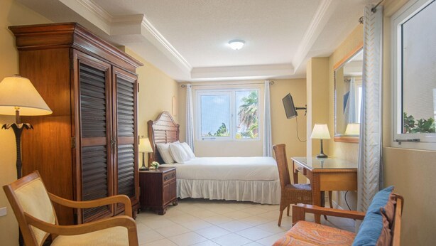 A hotel room with a bed, a desk, a chair, a wardrobe, and a window with a view of palm trees. - NEW Bay Harbor Studio 203 (Oranjestad)